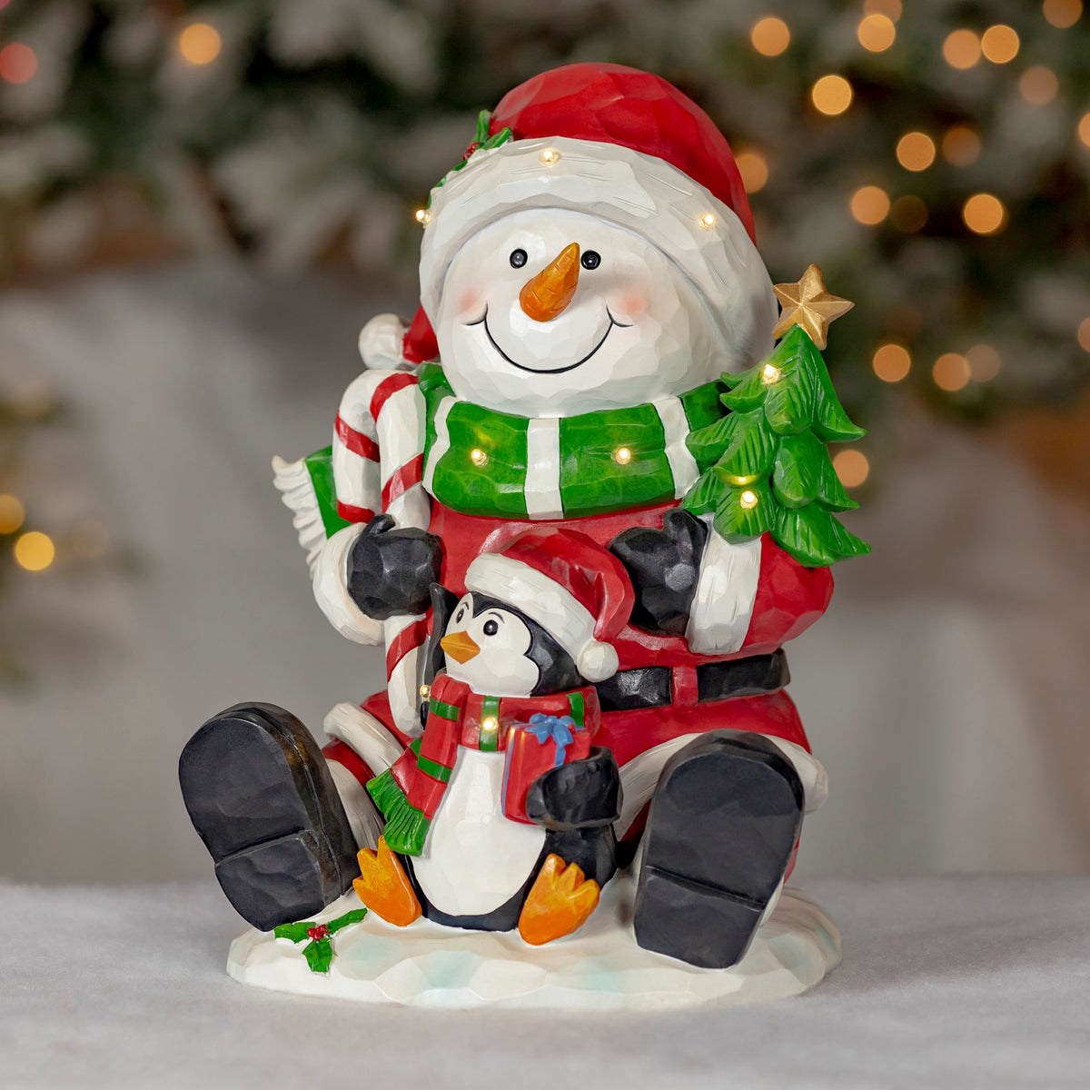 A smiling snowman in a Santa hat sits with a small penguin, a candy cane and a lit Christmas tree.