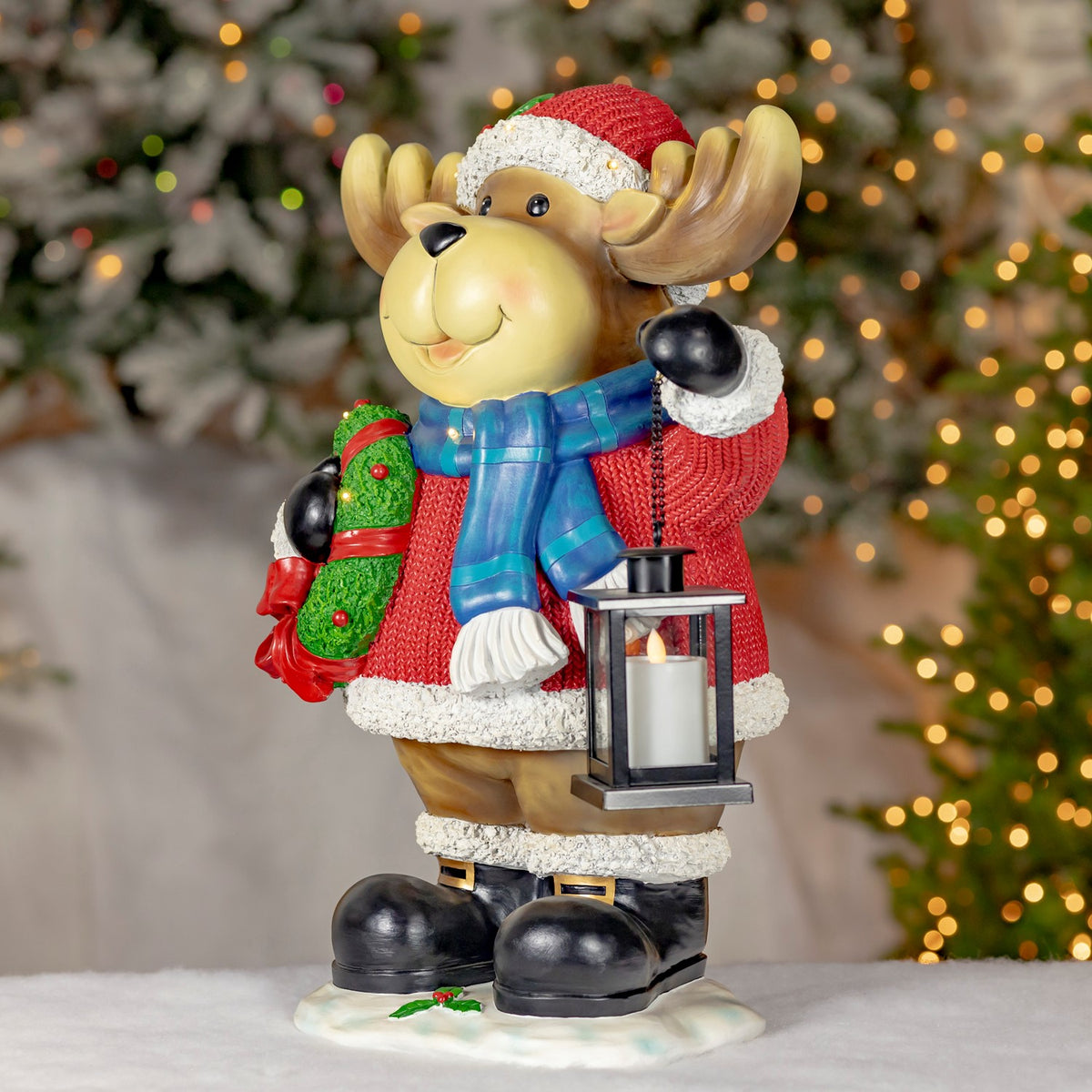 A full-body moose in a Santa hat holds a wreath and lantern against a softly lit Christmas tree.