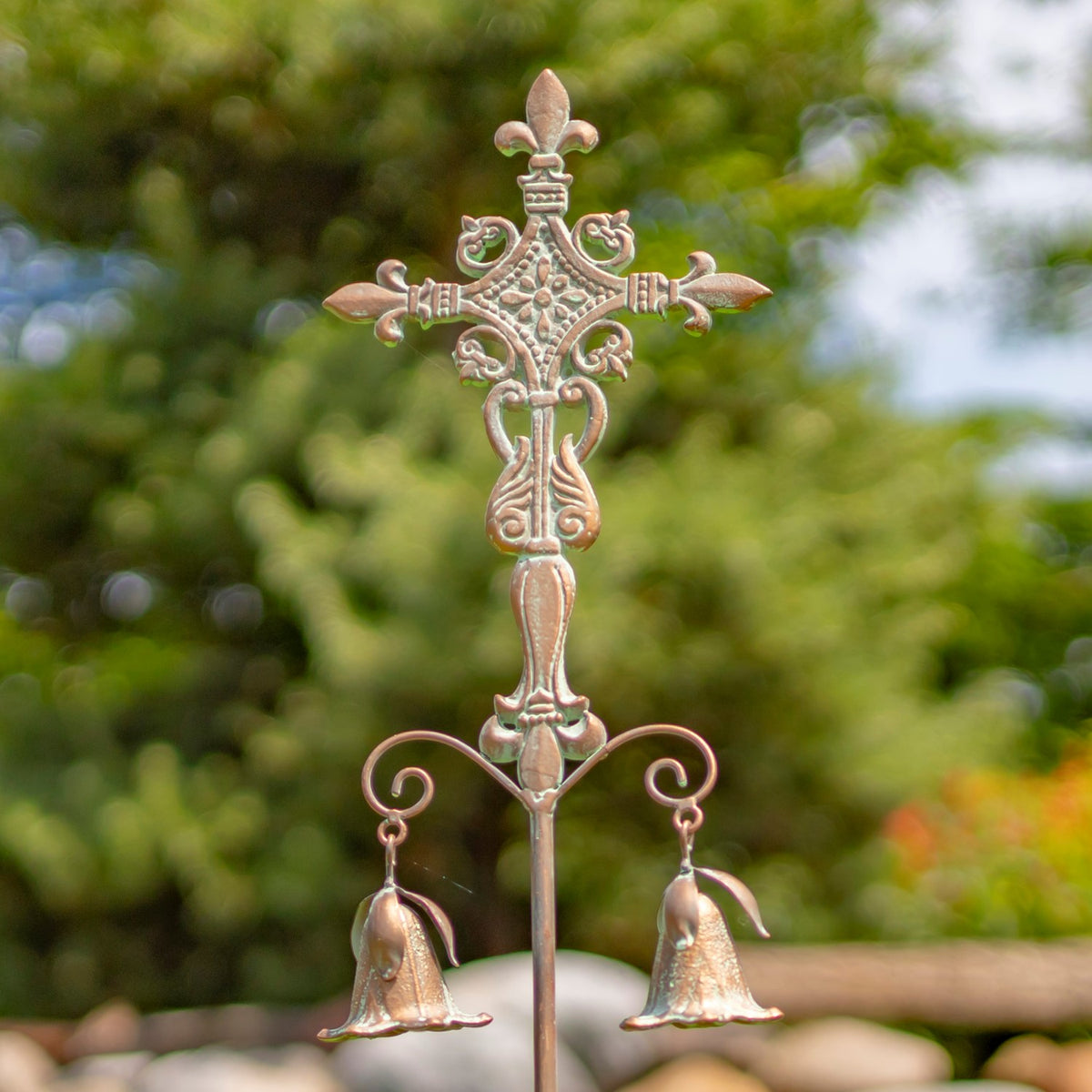 Close view of an ornate garden cross stake with two hanging bell-shaped ornaments against leafy greenery.