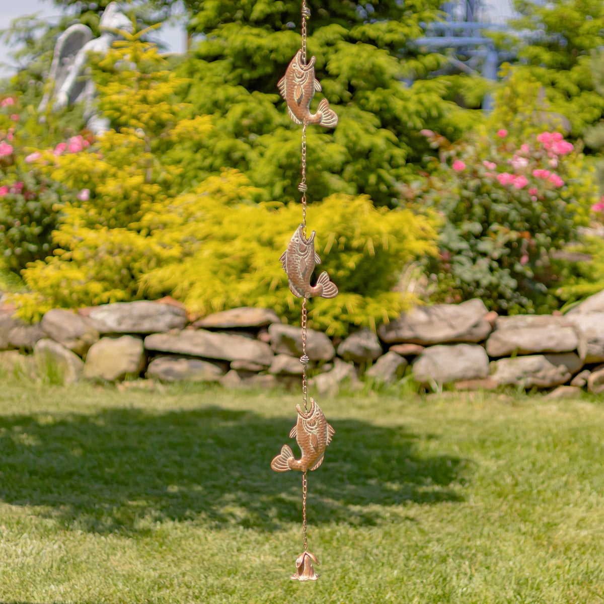 A copper-toned rain chain with jumping fish hangs over a lawn in a flower-filled garden.