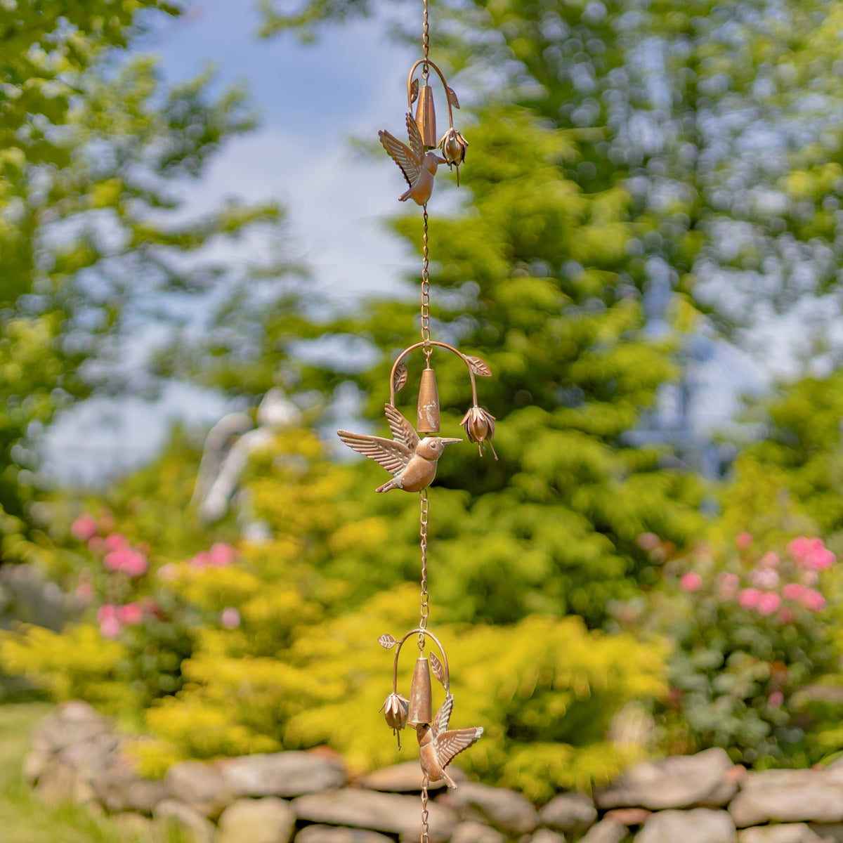 A hanging chain with three hummingbird ornaments and flower-shaped bells against a leafy garden.
