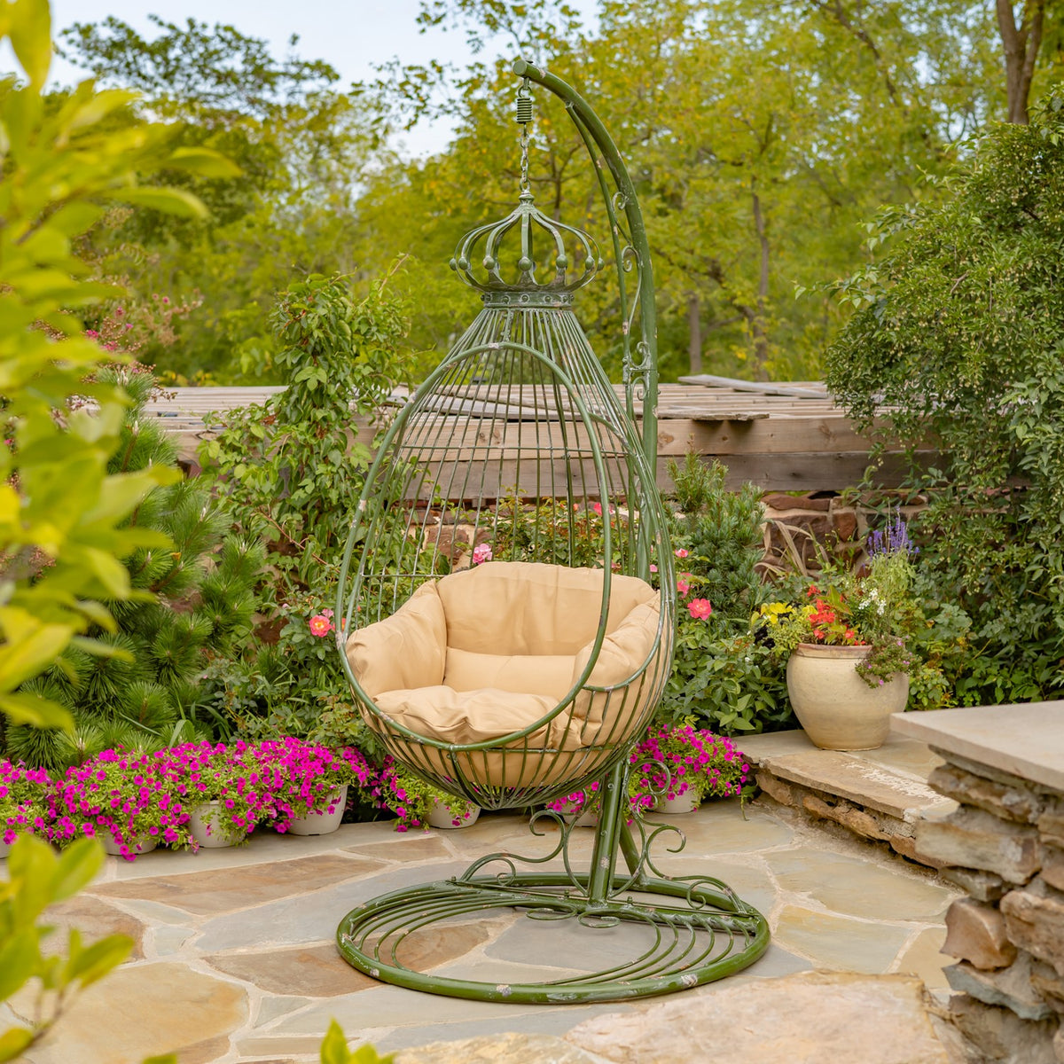 Green metal egg chair with a tan cushion and circular stand among garden flowers.