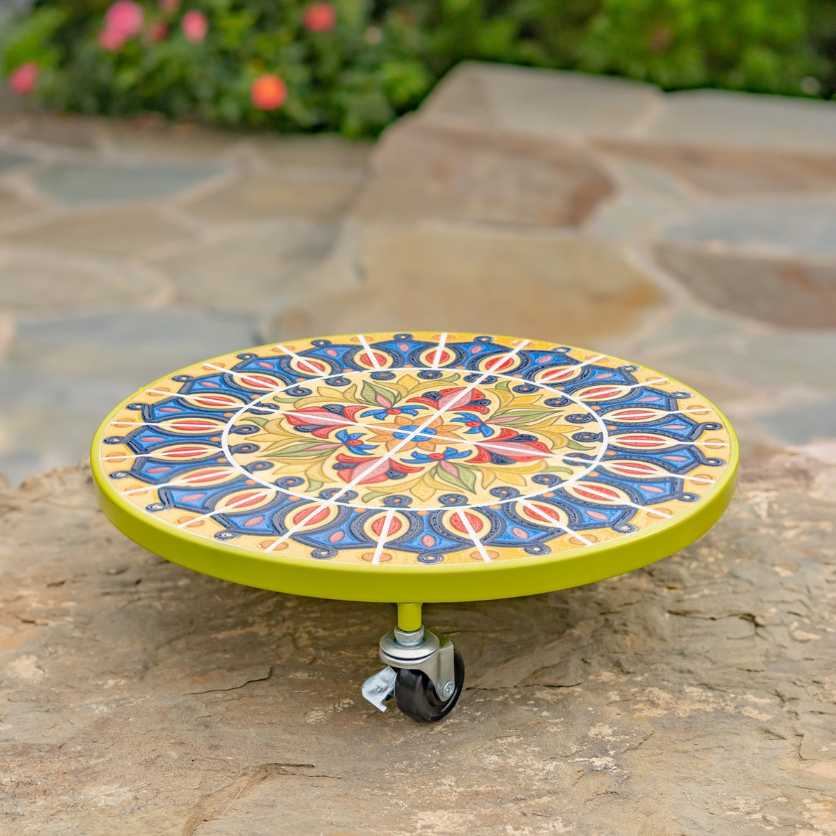 Round floral mosaic plant trolley with a lime-green rim and caster wheels on a stone path.
