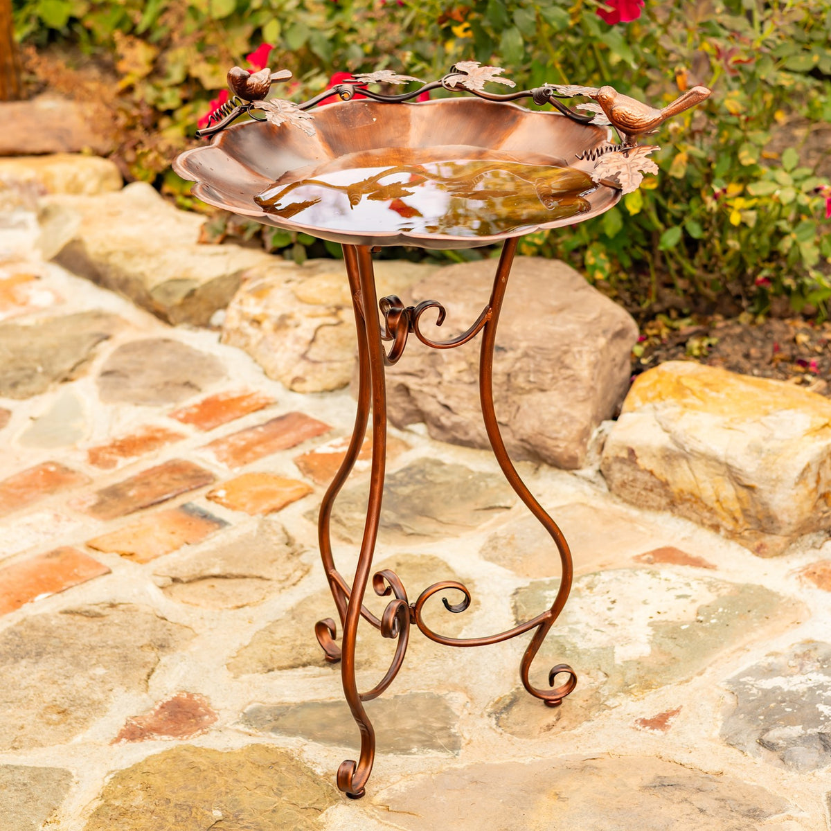 Flower-shaped birdbath on an ornate three-legged stand beside a stone path.