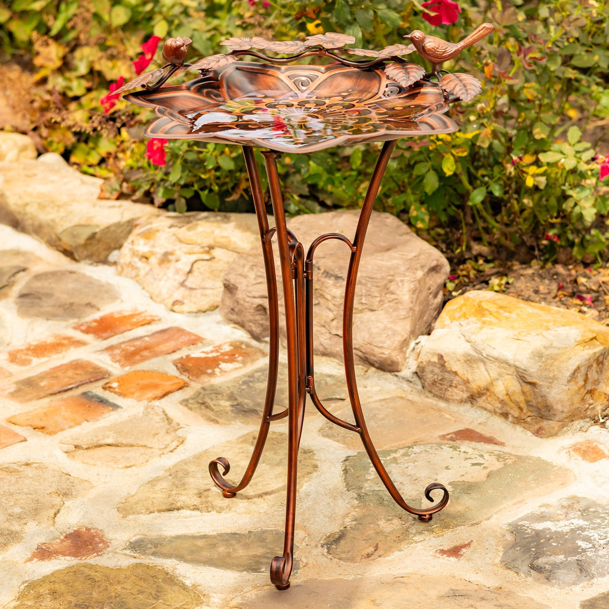 Bronze-toned birdbath on a curled three-legged stand, displayed on a stone patio among garden plants.