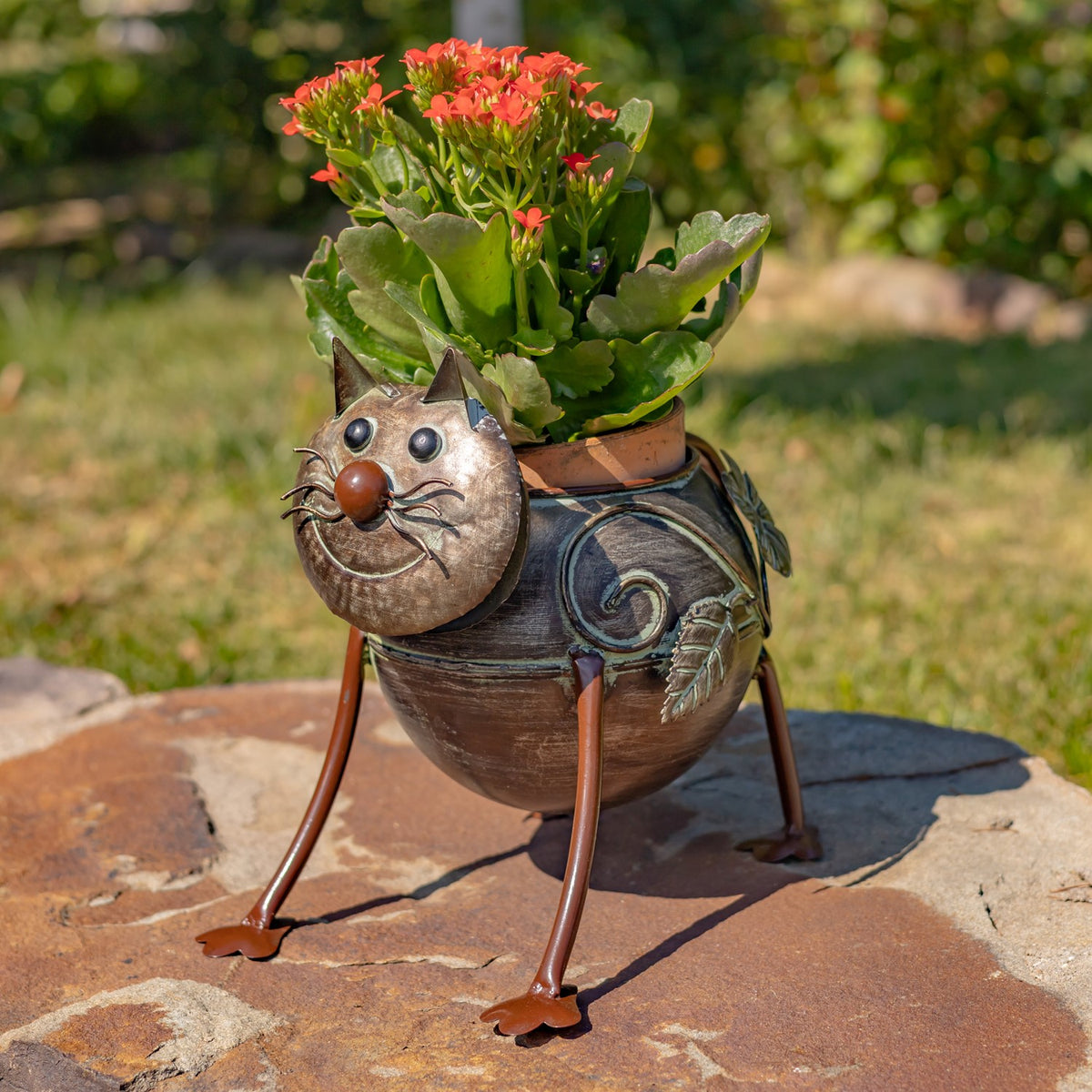 A cat-shaped metal planter on long legs holds green foliage and small red flowers outdoors.