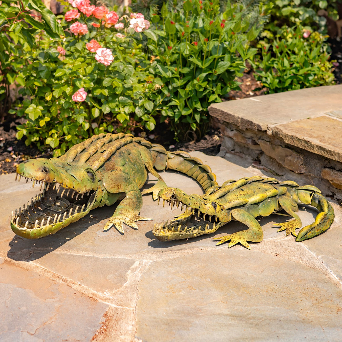 Two weathered green metal alligator decorations lie side by side on a stone patio among flowering plants.