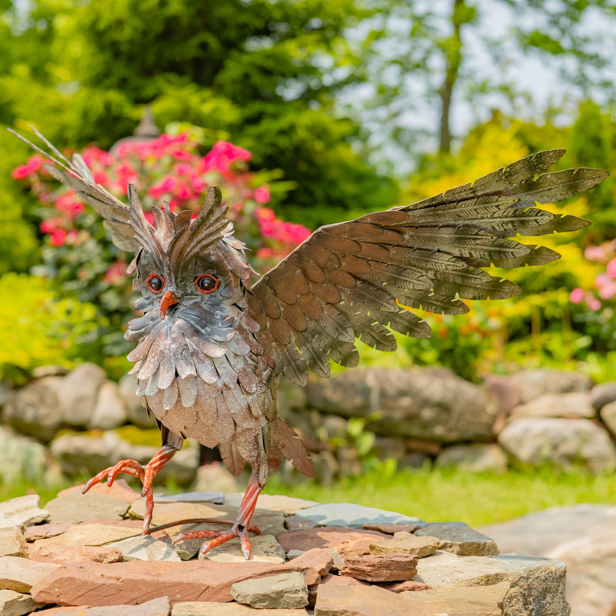Metal owl sculpture stands on a stone wall with one wing extended in a garden.