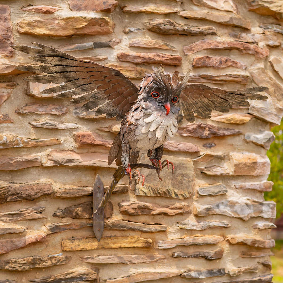 Metal owl sculpture with wings spread, perched on a branch against a stone wall.
