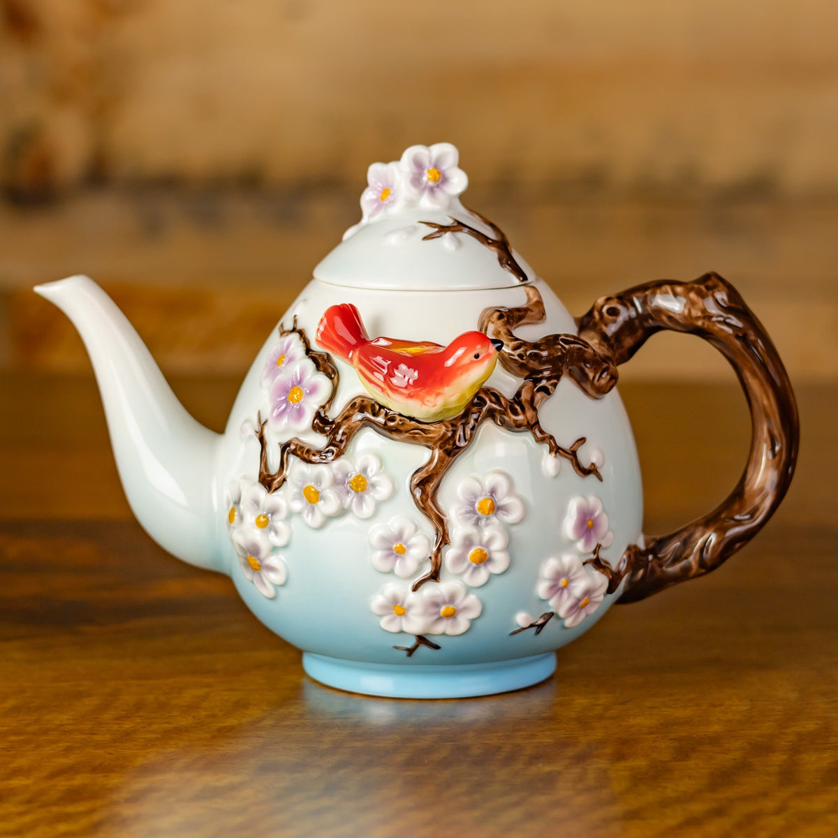 Decorative teapot with a red bird, raised brown branches, and pink and white flowers on a wooden table.