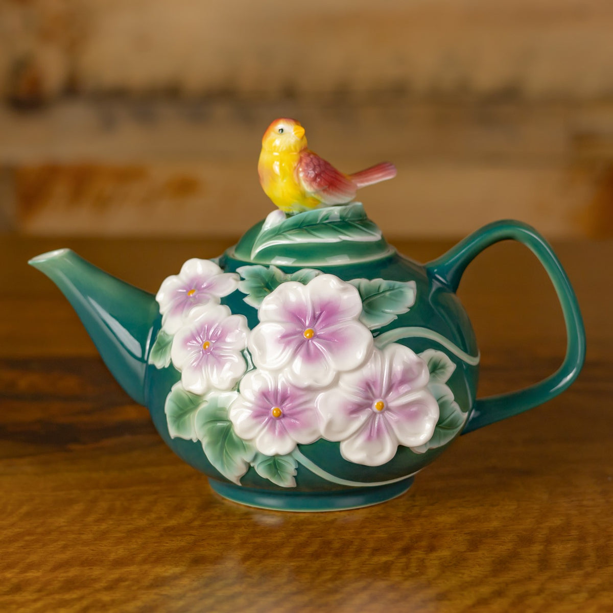 Teal decorative teapot with raised pink flowers and green leaves, topped by a small yellow bird.