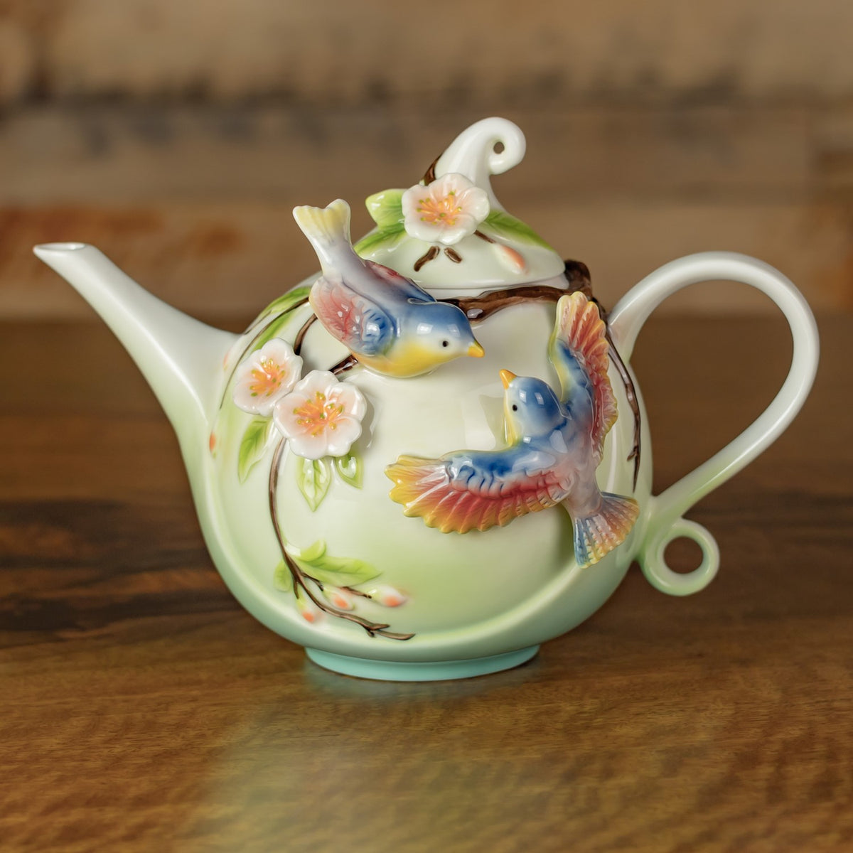 Pale green teapot with raised blue birds and pink flowers, shown from the spout side on a wooden surface.