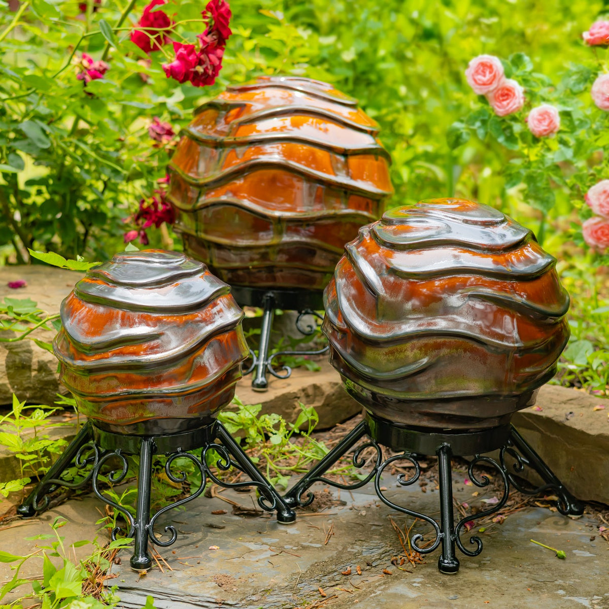 Three wavy, glossy orange and gray globes on black metal stands among garden flowers and greenery.