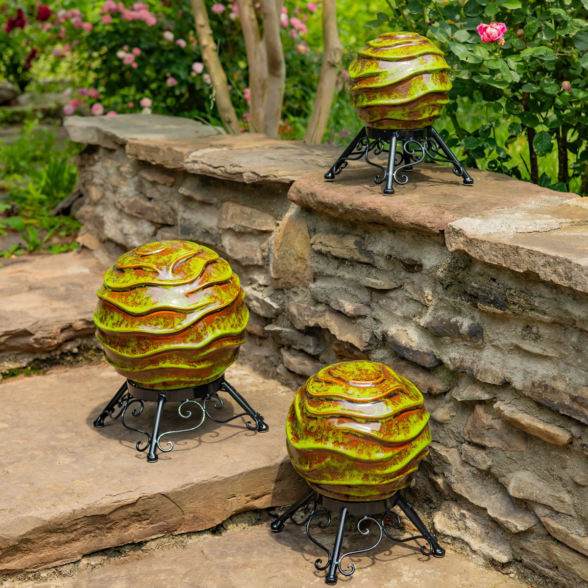 Three green and gold ridged gazing globes on black scrollwork stands among stone walls and flowering plants.
