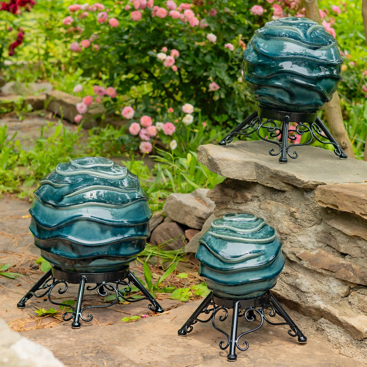 Three glossy teal, ridged garden globes on black scrollwork stands among flowers and stonework.