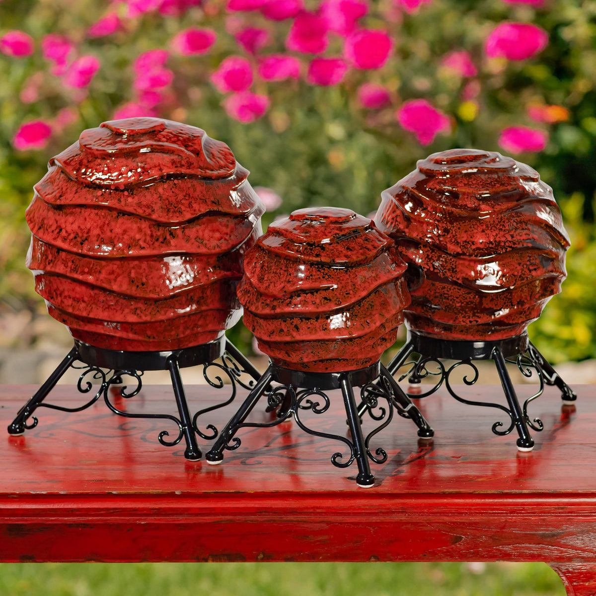 Three glossy red, ridged globes on black scrollwork stands displayed on a red table among flowers.