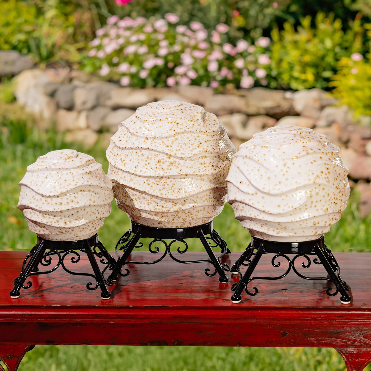 Three speckled ivory globes in black metal stands arranged on a red garden table.