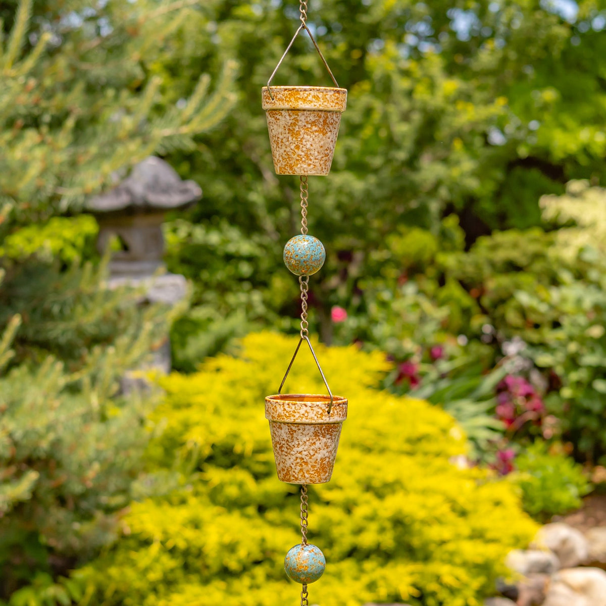 A hanging rain chain with speckled terracotta-colored cups and blue-green beads among garden greenery.