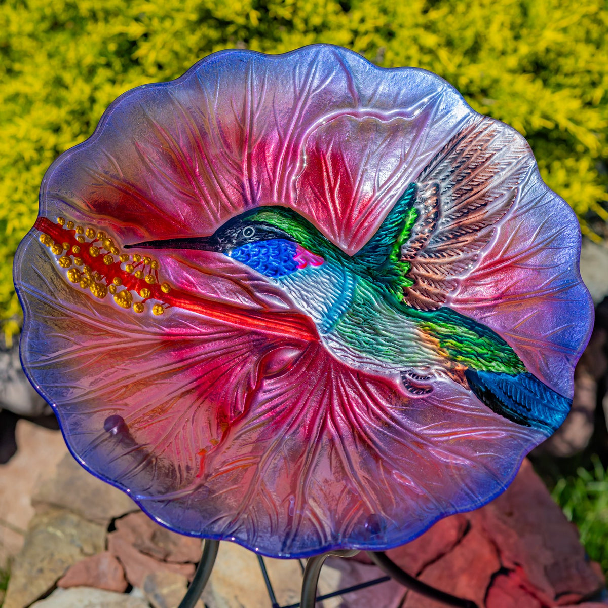 A pink and blue glass bowl with a raised hummingbird design, shown outdoors on its metal stand.