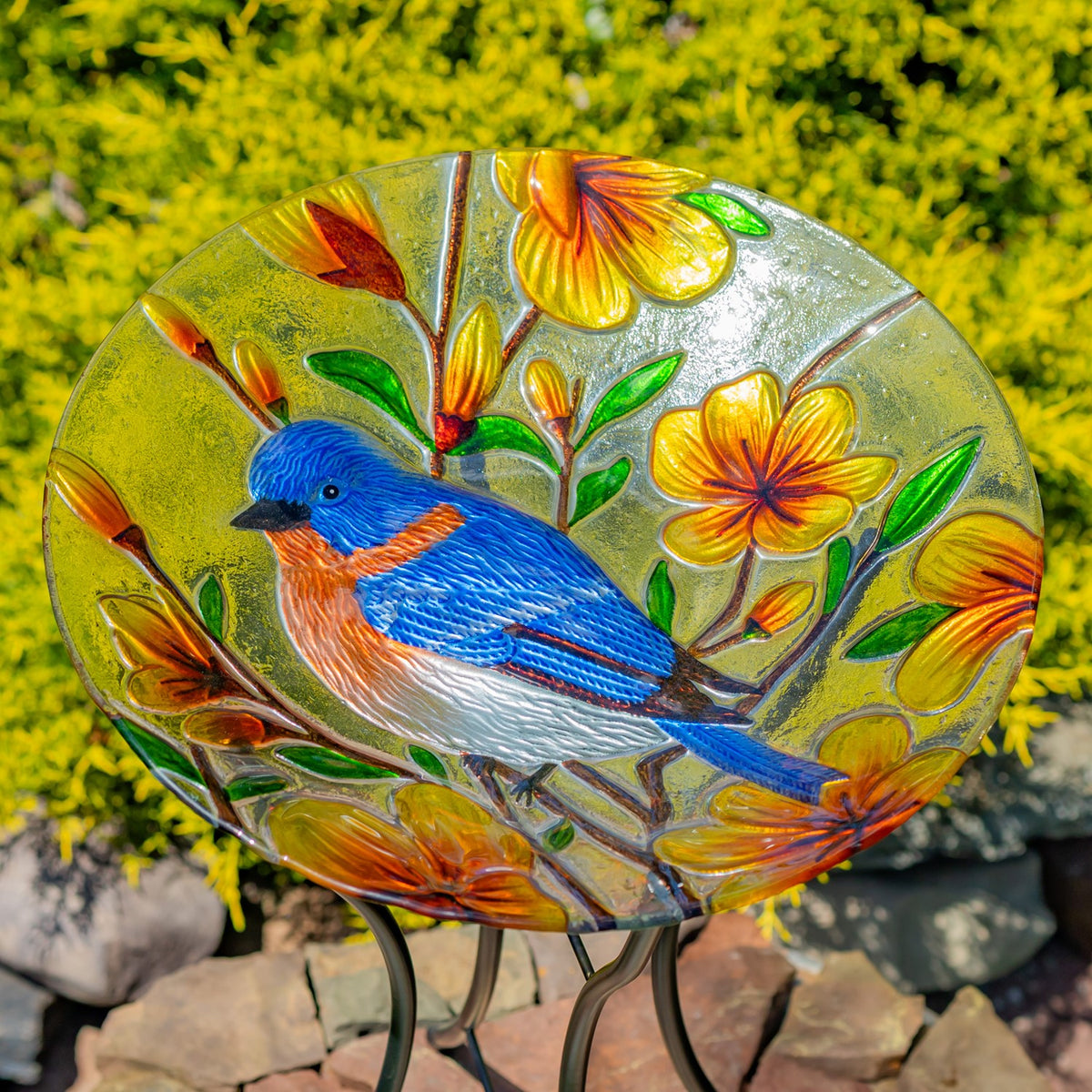 Colorful glass birdbath bowl with a bluebird and flowers, shown against garden greenery.