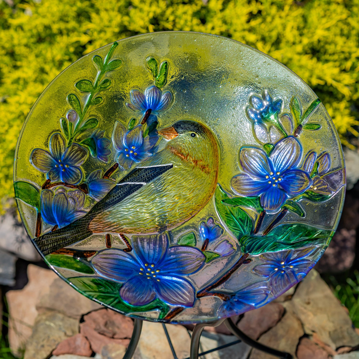 Colorful glass birdbath bowl with a goldfinch and blue flowers, shown upright on its metal stand outdoors.