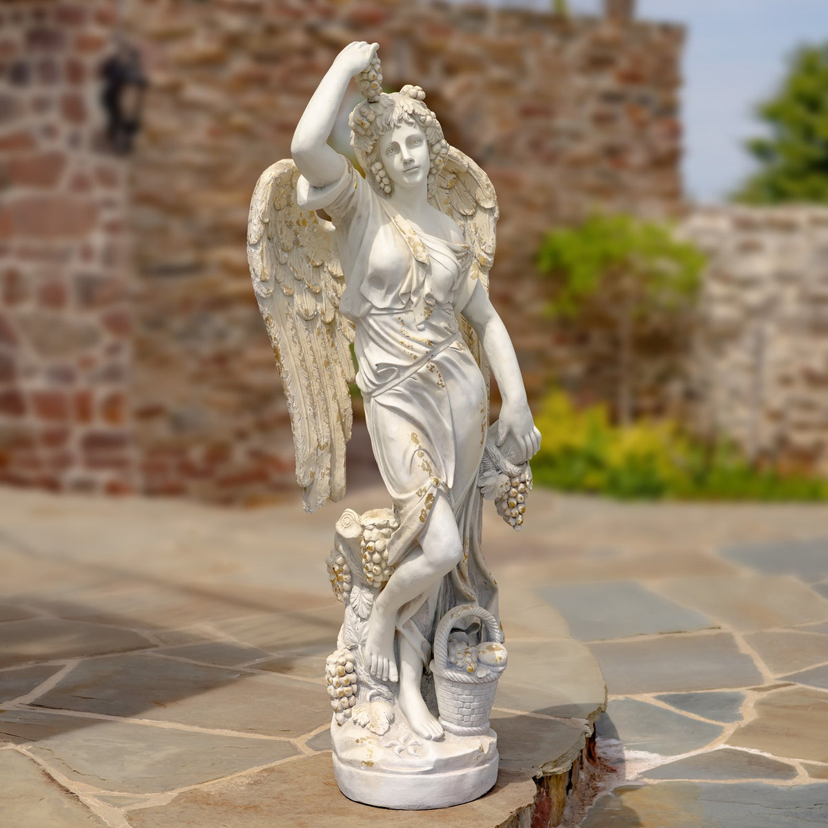 Full view of a pale angel statue with raised arm, wings, draped clothing, grapes and a basket on a stone patio.