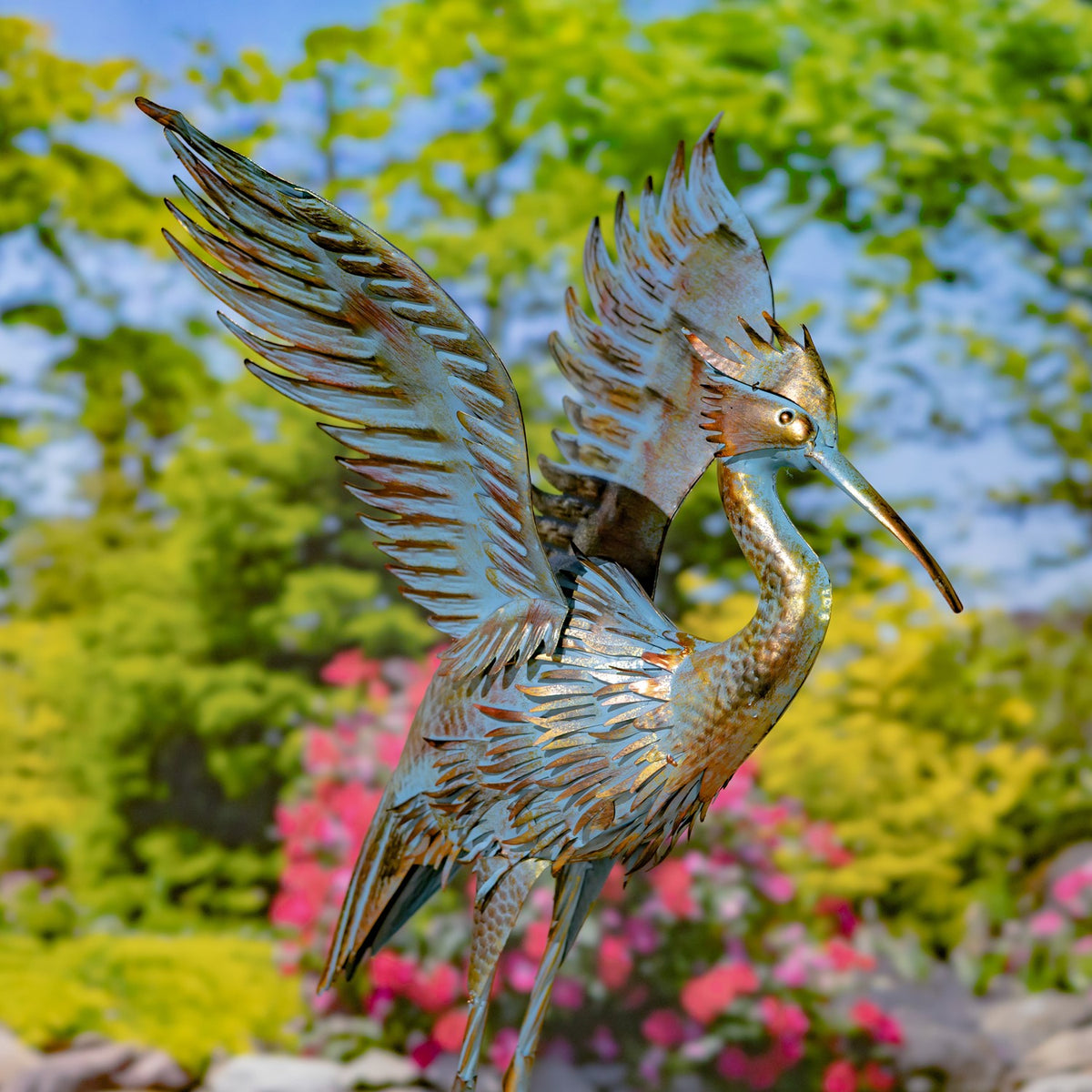 Metal heron garden sculpture with wings raised against flowering shrubs and greenery.