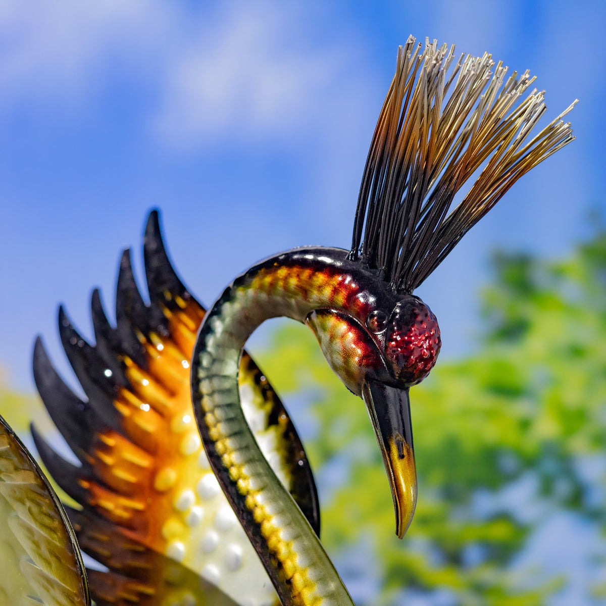 Close-up of a metallic heron figurine’s curved neck, long beak, and feathered crest against a garden backdrop.