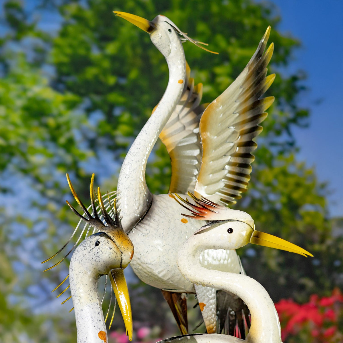 Close view of white metal heron figurines with raised wings and yellow beaks against garden greenery.