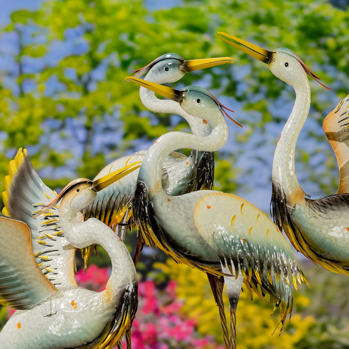 Close view of several painted metal crane figurines with long bills and feathered wings in a garden setting.