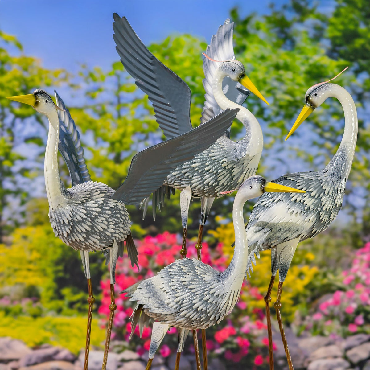 Four metal heron garden figures in varied poses stand among flowers and greenery.