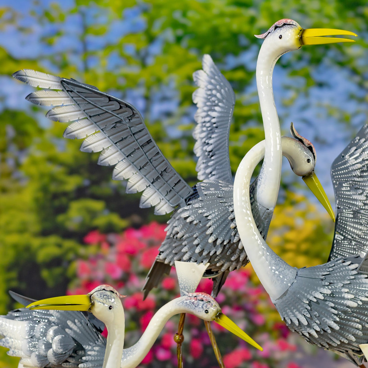 Several gray-and-white metal heron garden figures with raised wings and yellow beaks against a flower-filled garden.