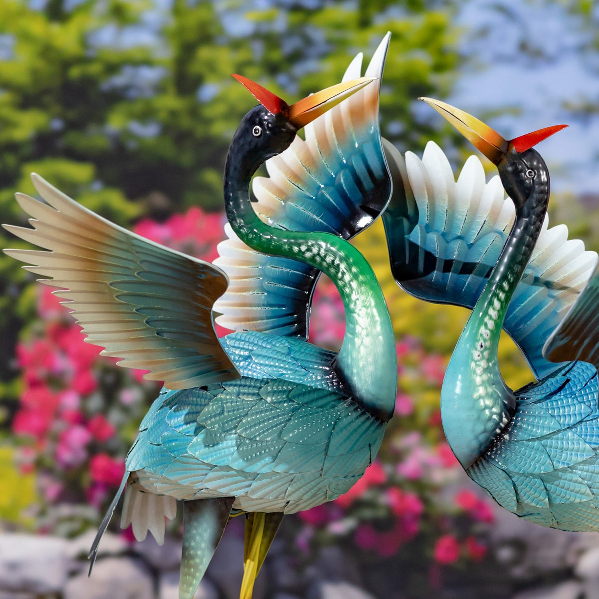 Two blue heron garden figurines with raised wings and red head feathers, shown among flowers.