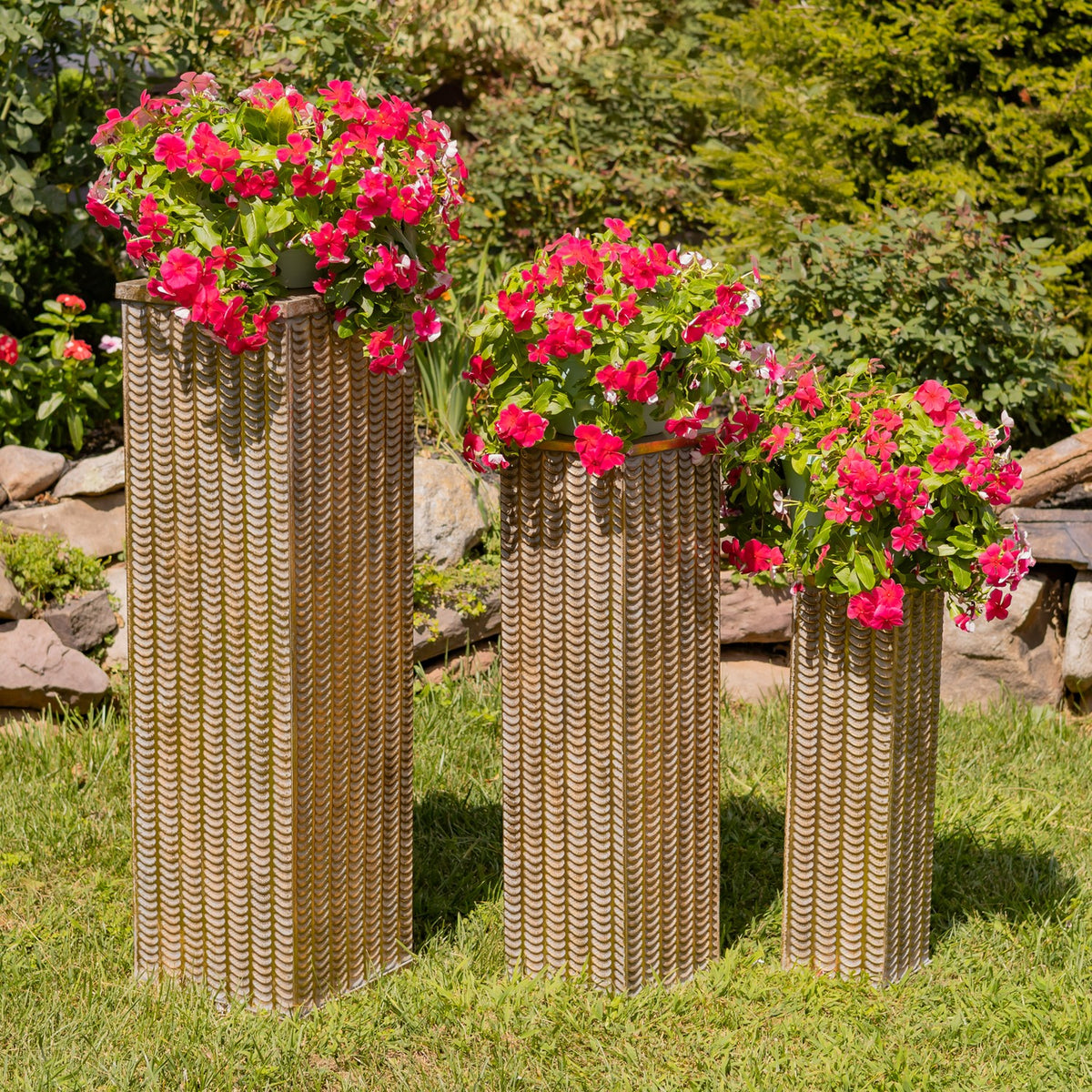 Three textured gold-toned pedestal pillars stand on grass, topped with bright pink flowers.
