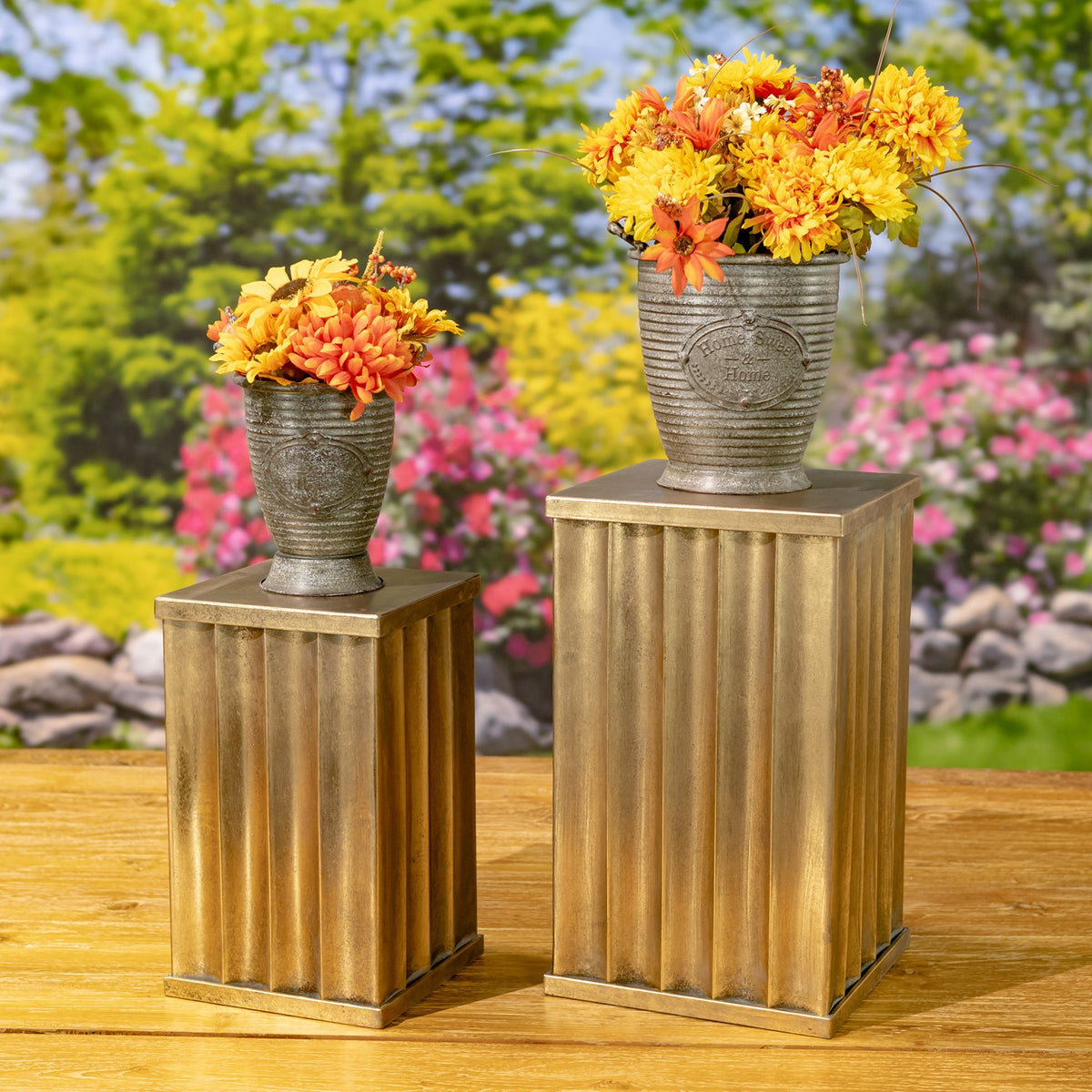 Two fluted gold-toned pedestal pillars display flower-filled pots in a garden setting.