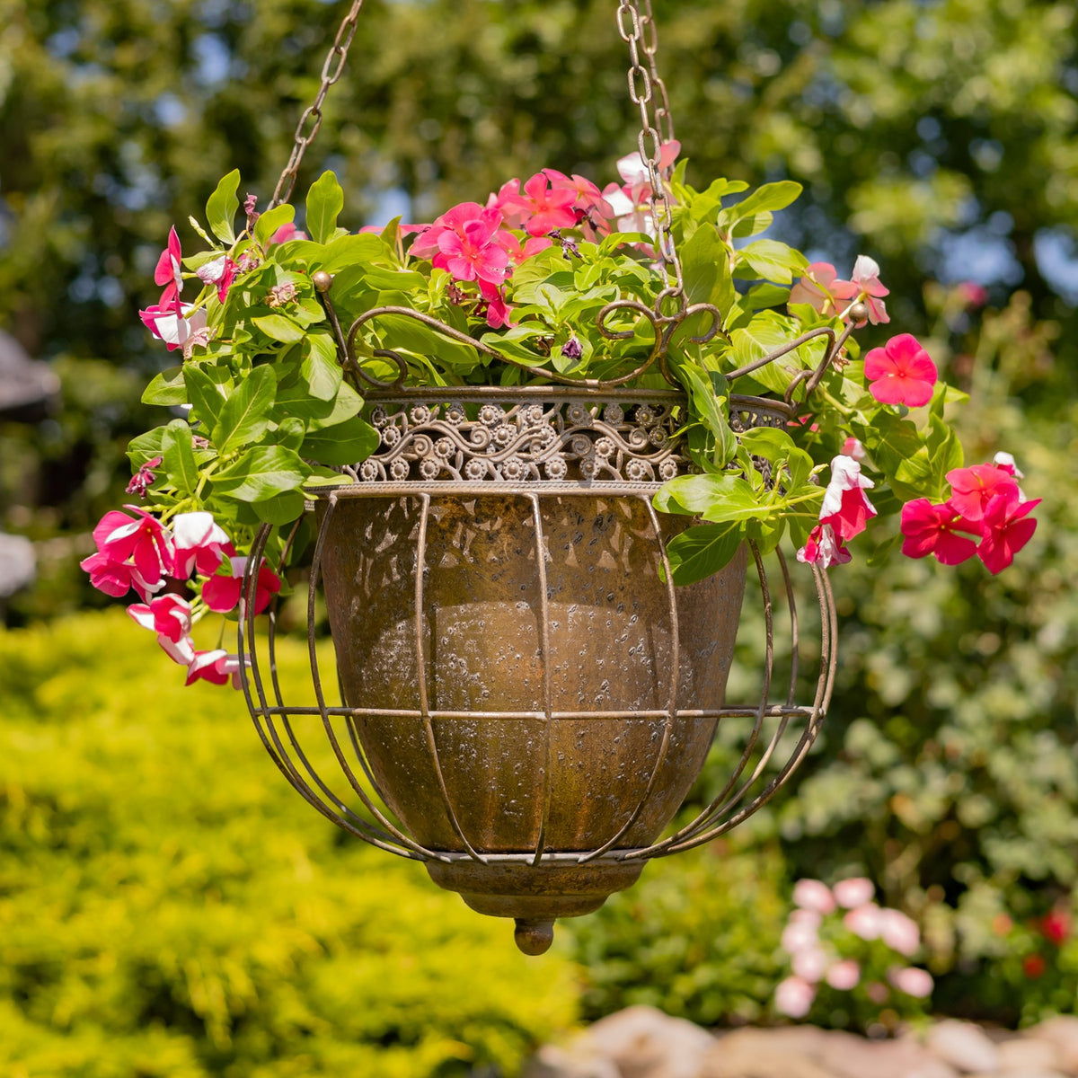 Hanging metal basket planter with a brown inner pot, pink blooms, and trailing greenery outdoors.