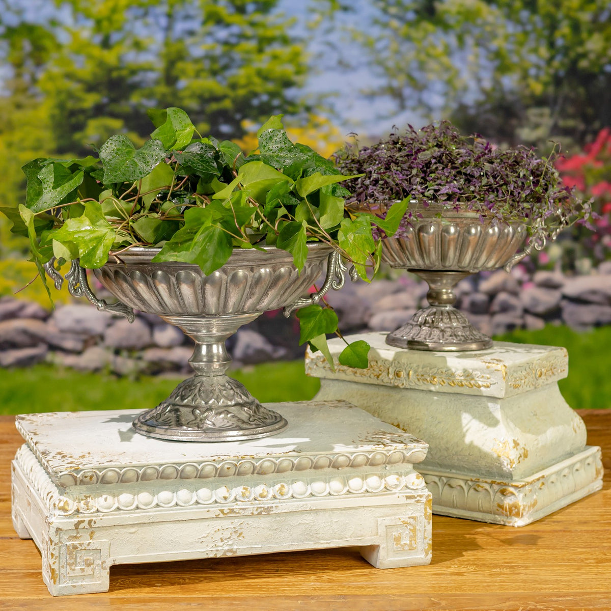 Two ornate silver urn planters filled with greenery sit on decorative cream pedestals.