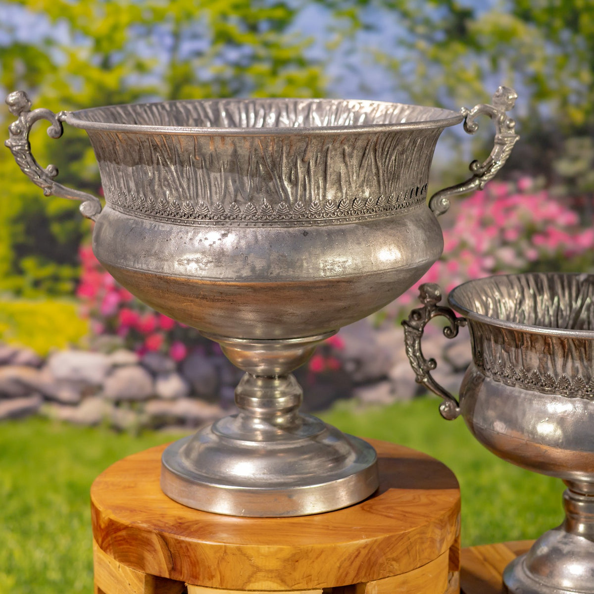 Large ornate silver urn with curled handles on a wooden pedestal, with a smaller urn partly visible beside it.