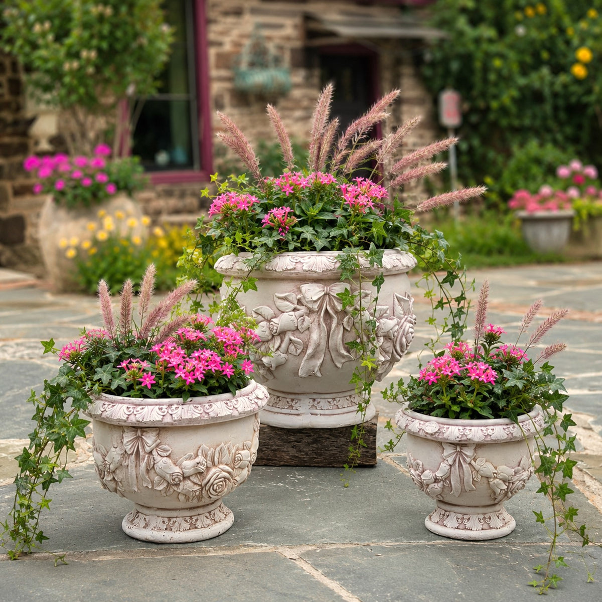 Three ornate pale urn planters filled with pink flowers and trailing greenery on a garden patio.