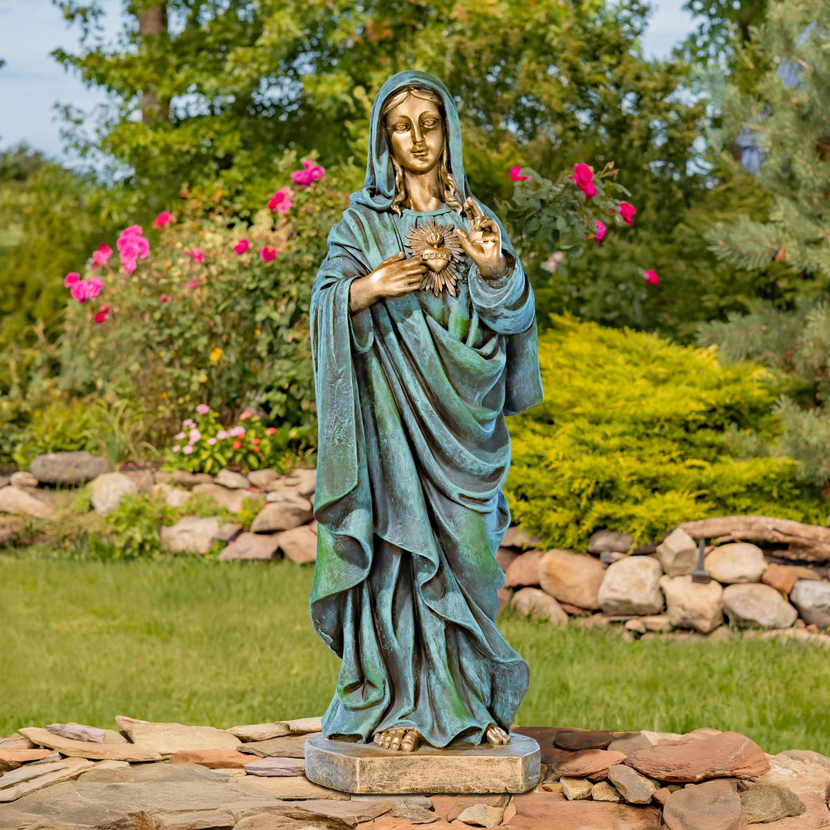 Full-length statue of Mary with a radiant heart stands on a stone base among garden flowers.