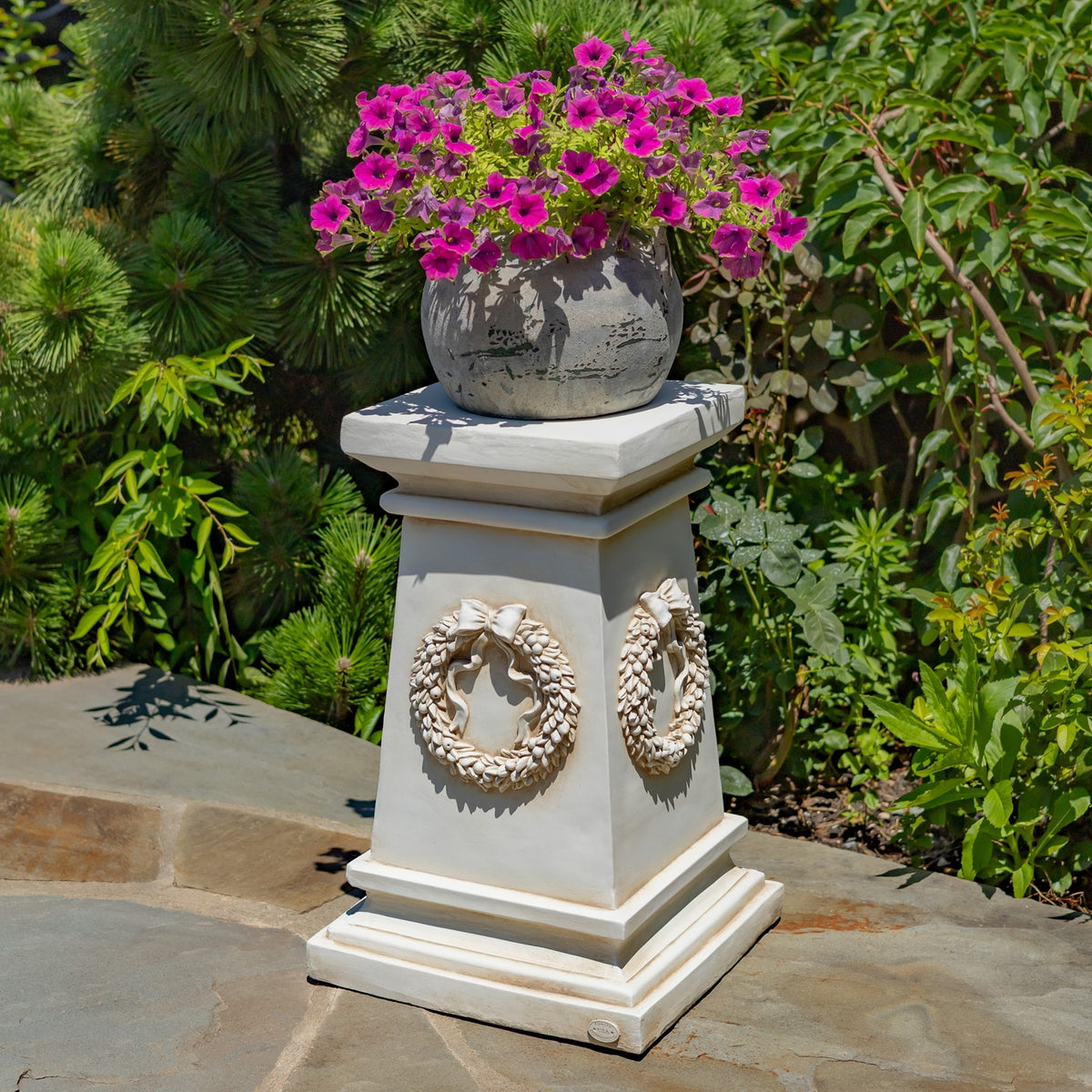 Light-colored decorative column with wreath reliefs supporting a flowerpot of bright pink blooms in a garden.
