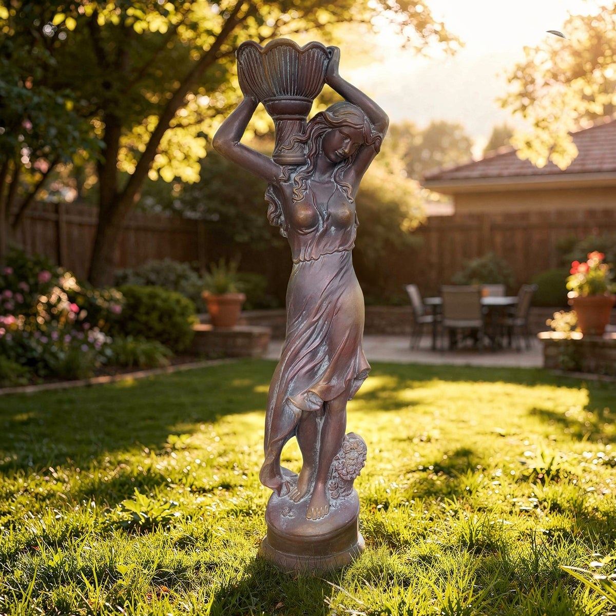 A full-length statue of a woman holding a flower pot overhead stands on a lawn in a sunlit garden.