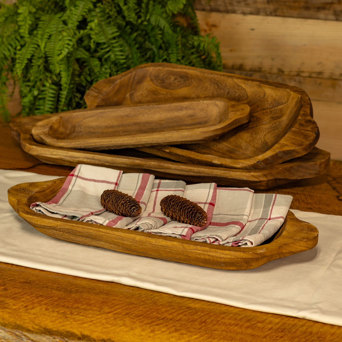 Four wooden trays are stacked and arranged with folded plaid cloths and two pinecones.