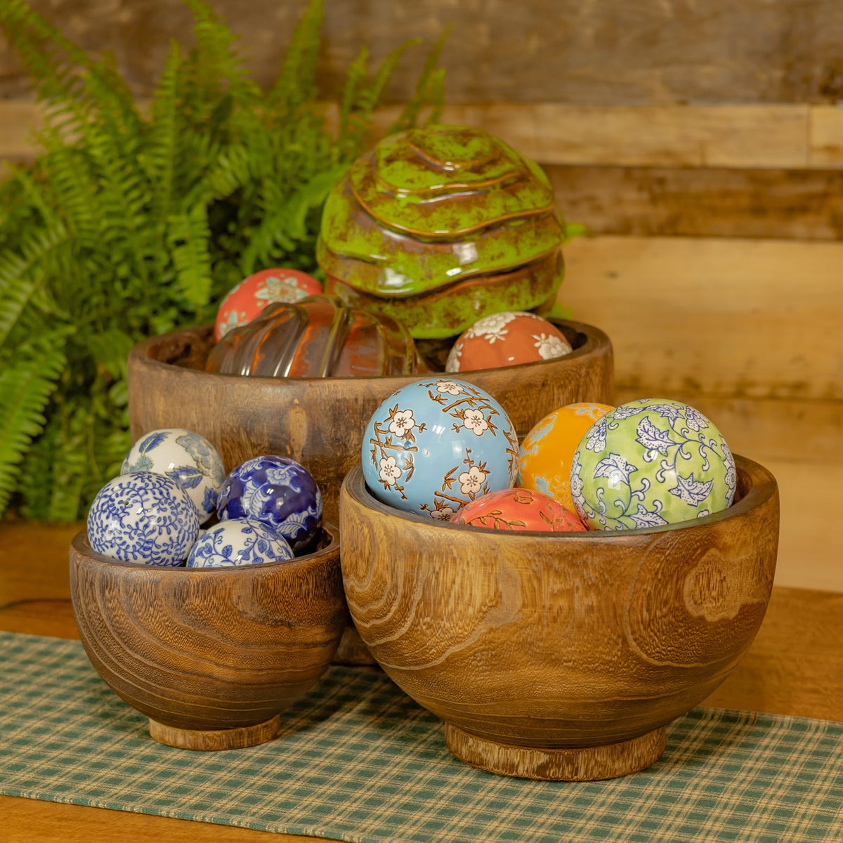 Three round wooden bowls hold colorful patterned ornaments on a table beside a fern.