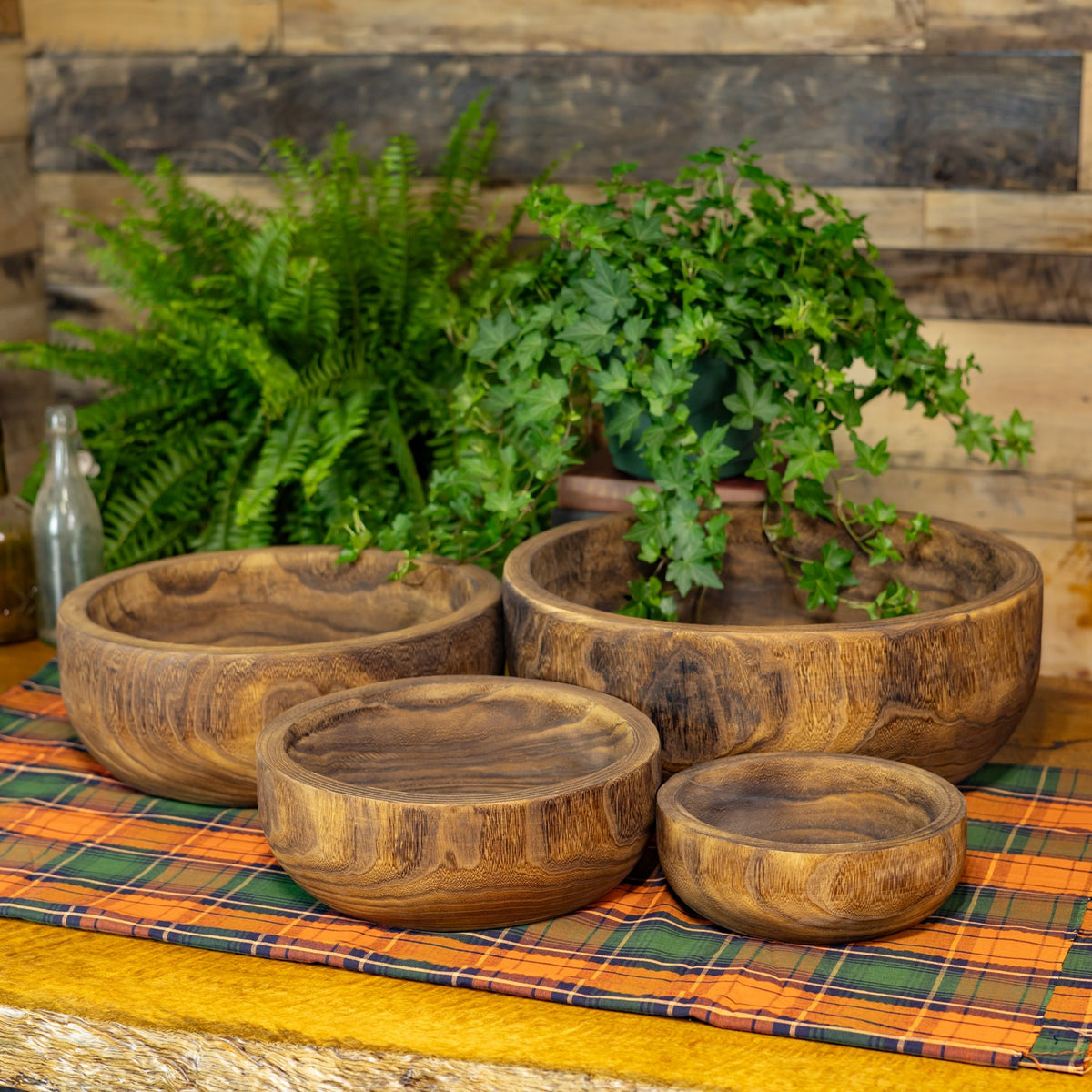 Four round wooden bowls in different sizes arranged on a plaid cloth, with leafy plants behind them.