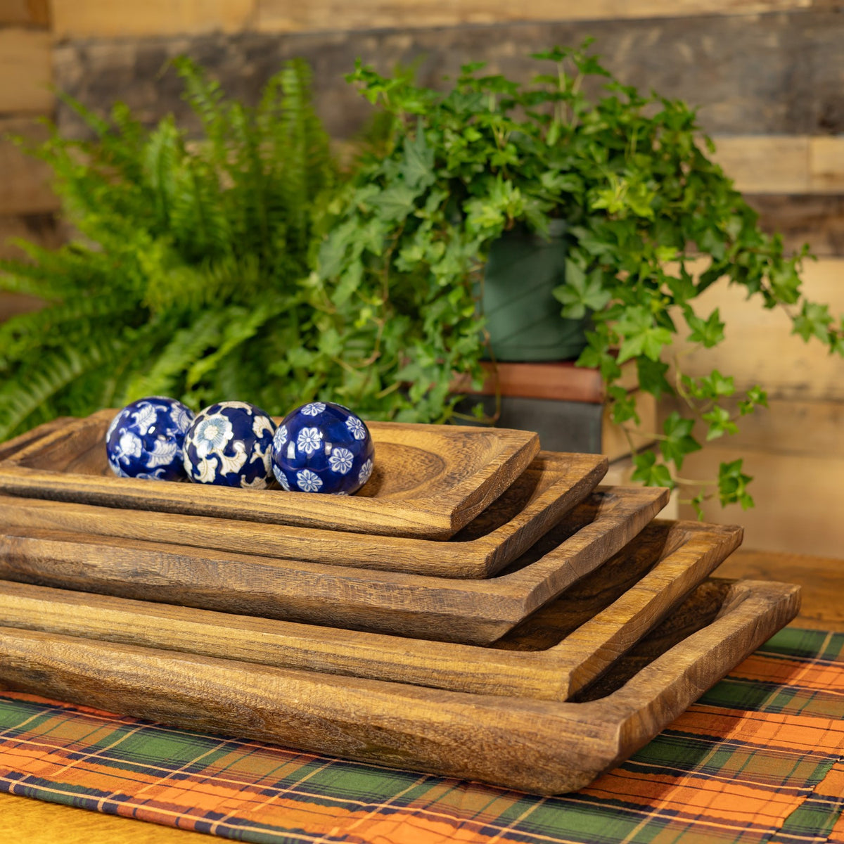 Five rustic wooden trays nested on a plaid cloth, with blue-and-white decorative balls on top.