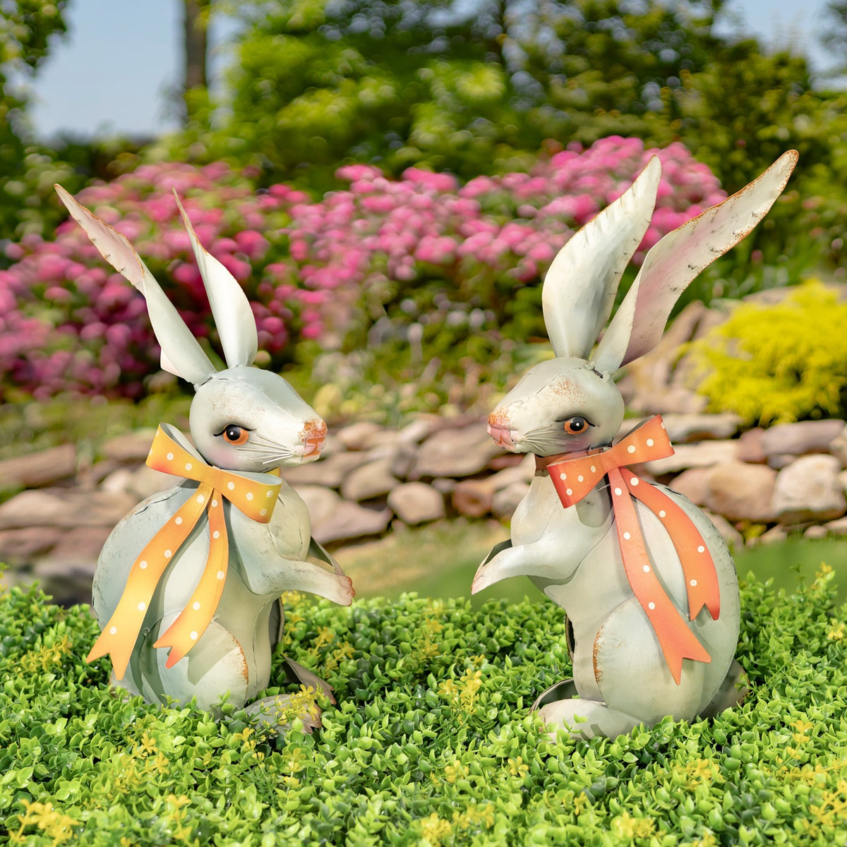 Two pale metal rabbit figurines with orange polka-dot ribbons sit among green ground cover and garden flowers.