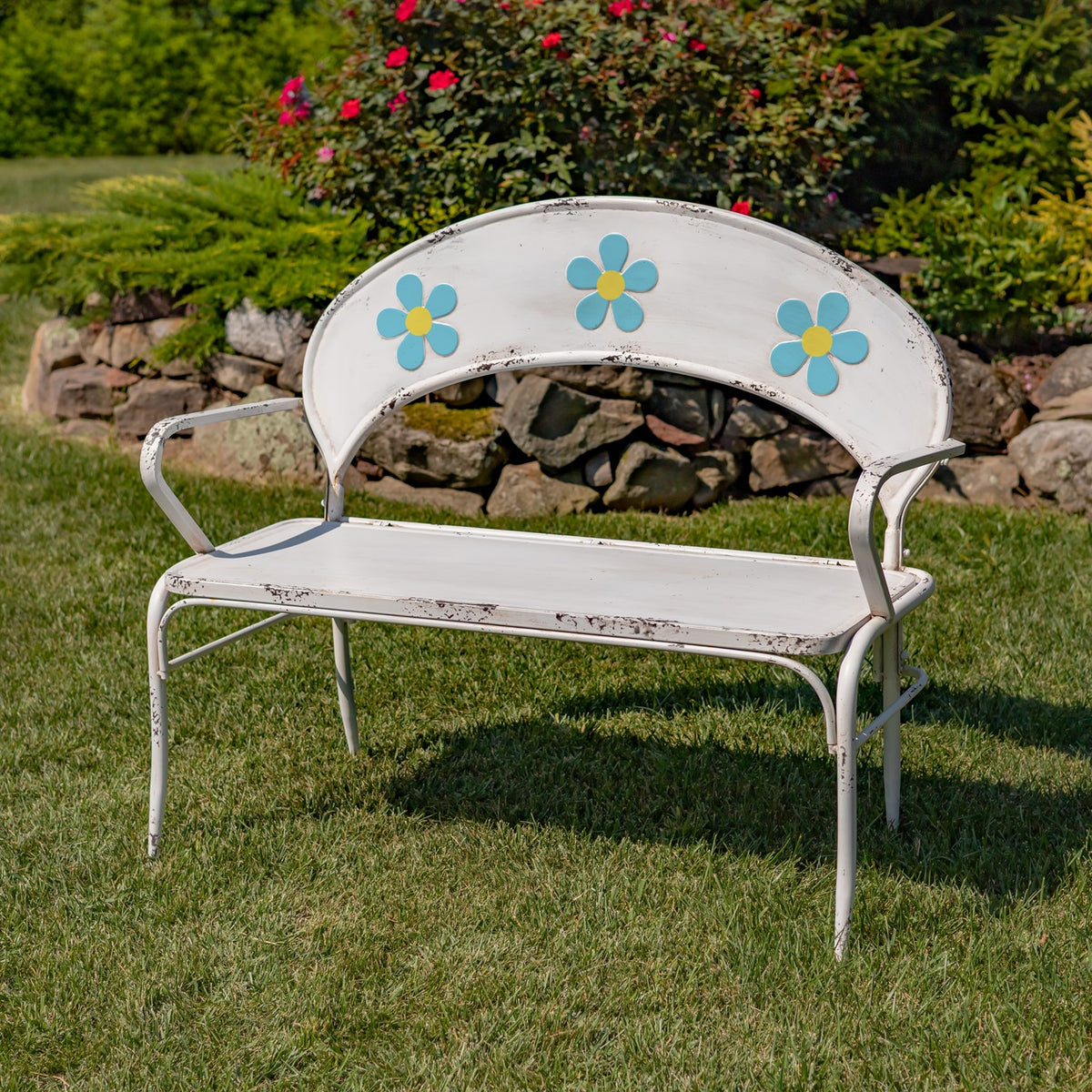 White metal garden bench with blue and yellow flower accents on a lawn beside a stone border.