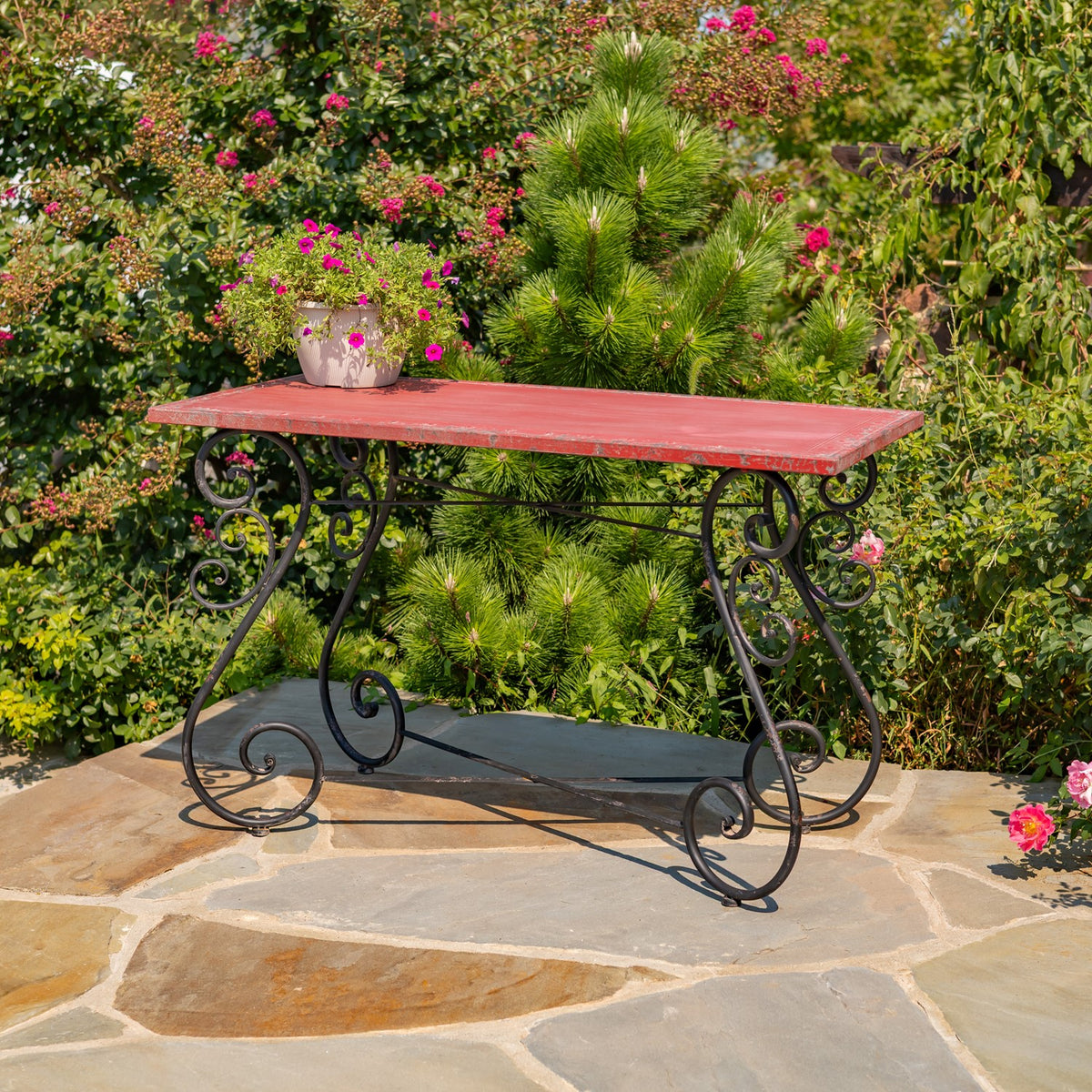 Red rectangular tabletop on dark curled iron legs, photographed on a stone patio against garden greenery.