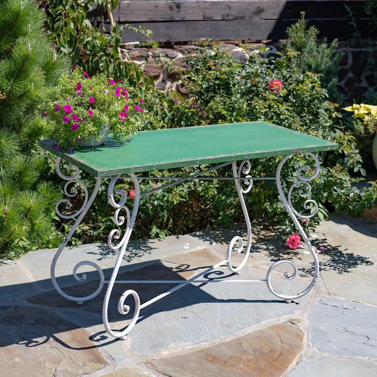 Green rectangular table with pale scrollwork legs on a stone patio among flowering garden plants.